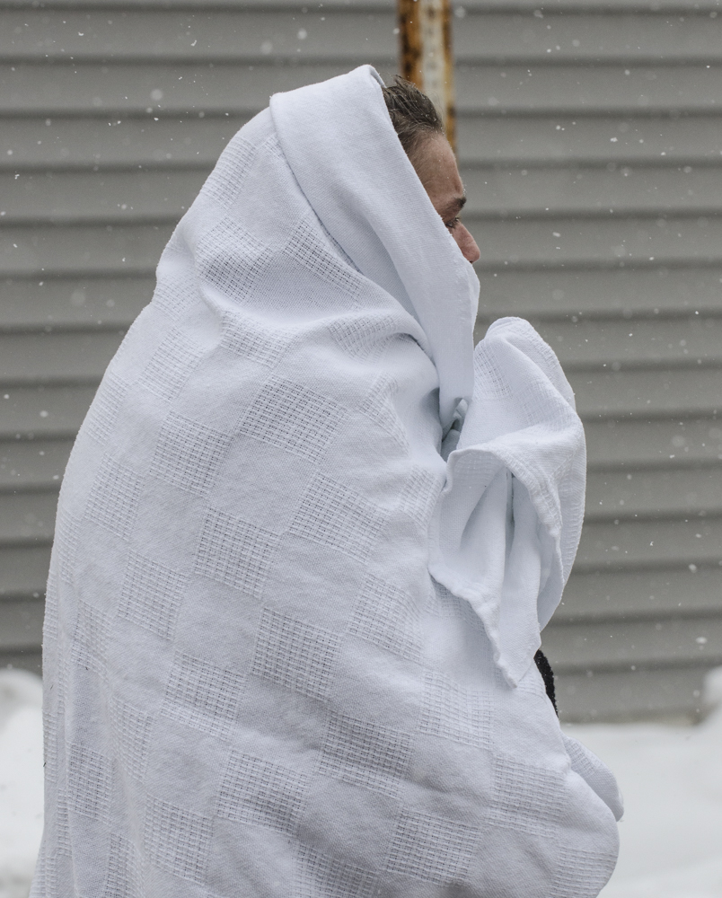 A resident stands outside the apartment, wrapped in a thin blanket against the chill and snowfall.